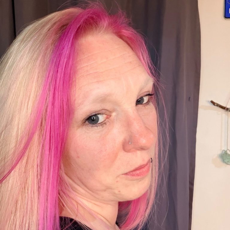 Frau mit langen, hellblonden Haaren und pinken Strähnen, schaut über die Schulter.